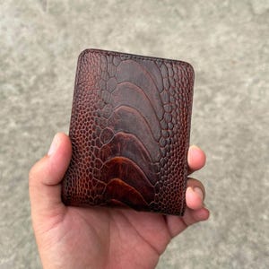 Pode incluir: Uma carteira dobrável castanho-escura com um padrão texturizado distinto que lembra pele de avestruz. A carteira tem uma forma retangular com cantos arredondados e uma cor mogno profunda e rica com detalhes em castanho mais escuro.