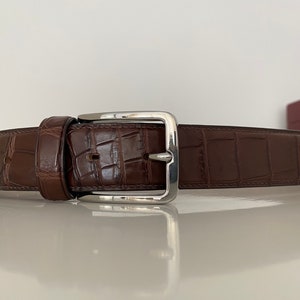 Op de afbeelding: Een bruine leren riem met een zilveren gesp. De riem is gemaakt van alligatorshuid en heeft een klassiek ontwerp.