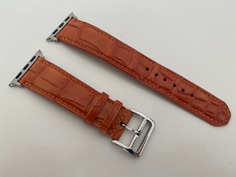 May include: A brown leather watch band with a silver buckle. The band has a crocodile pattern and is stitched with a contrasting orange thread.