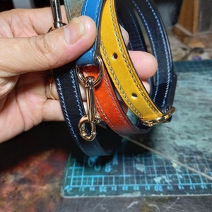 May include: A selection of colourful leather straps with gold-coloured metal clasps. The straps are in shades of blue, orange, and yellow, with contrasting stitching. The leather appears to be high-quality, and the clasps are shiny.