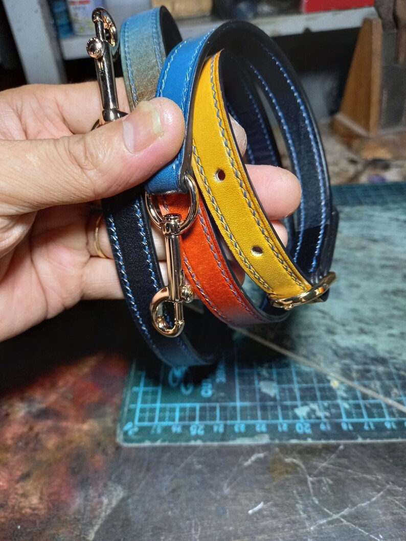 May include: A selection of colourful leather straps with gold-coloured metal clasps. The straps are in shades of blue, orange, and yellow, with contrasting stitching. The leather appears to be high-quality, and the clasps are shiny.
