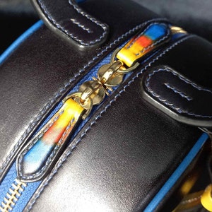 May include: Close-up of a black leather handbag with a blue zip and colourful zip pulls. The zip pulls have a gradient of yellow, orange, and blue. The bag has blue stitching and gold-coloured hardware.