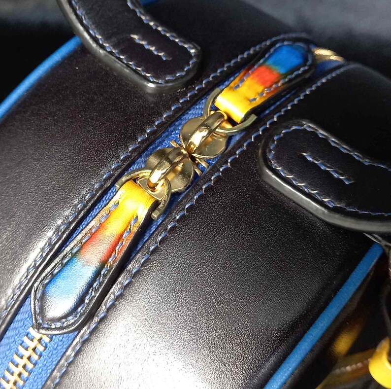 May include: Close-up of a black leather handbag with a blue zip and colourful zip pulls. The zip pulls have a gradient of yellow, orange, and blue. The bag has blue stitching and gold-coloured hardware.