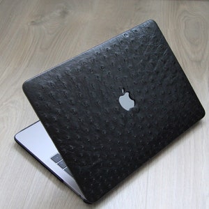 Könnte beinhalten: Ein schwarzer Laptop mit einer Hülle aus Lederimitat mit Straußenhautstruktur. Die Hülle hat ein silbernes Apple-Logo.