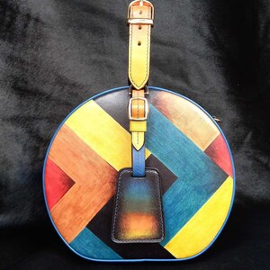 May include: A circular handbag featuring a vibrant geometric design in yellow, orange, red, blue, and black. It has a yellow leather handle with gold buckles and a blue luggage tag.
