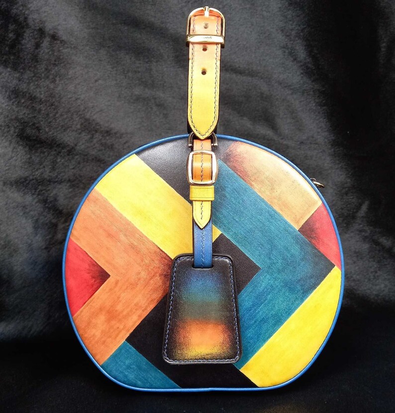 May include: A circular handbag featuring a vibrant geometric design in yellow, orange, red, blue, and black. It has a yellow leather handle with gold buckles and a blue luggage tag.
