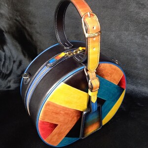 May include: A circular leather handbag in black with a geometric pattern in shades of blue, yellow, orange, and red. It has a top handle with a buckle, a blue zip, and a small tag. The bag's measurements are not visible.