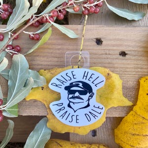 Könnte beinhalten: Ein weißer Aufkleber mit dem Text "RAISE HELL PRAISE DALE" und einer Schwarz-Weiß-Illustration eines Mannes mit Sonnenbrille und Baseballkappe. Der Aufkleber befindet sich auf einem gelben Blatt, mit Ästen und Beeren im Hintergrund.