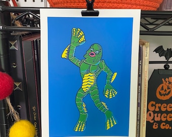 May include: A print featuring a green creature with yellow accents against a blue background. The creature has webbed hands and feet. The print is held by a black clip. A decorative orange pumpkin basket is in the background.