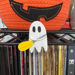 Może przedstawiać: Biała figurka ducha trzymająca żółtą pałkę do baseballu. Duch się uśmiecha i ma dwa czarne oczy. Figurka stoi na półce z różnymi książkami i płytami za nią.