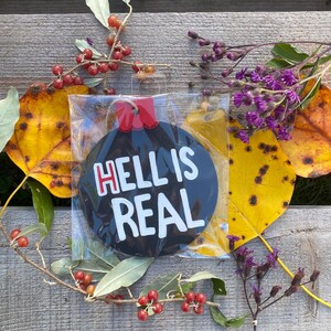 May include: A black and white ornament with the text "HELL IS REAL" in a clear plastic package. The ornament has a red top and a string for hanging. The ornament is surrounded by fall foliage, including yellow leaves and red berries.