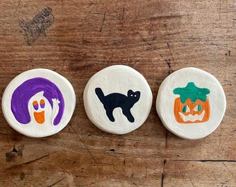 May include: Three round, white ceramic coasters with Halloween-themed designs. One features a purple-haired ghost with an orange face, another a black cat, and the last a jack-o'-lantern with a green stem. The coasters are arranged on a wooden surface.