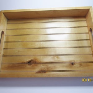 May include: A wooden serving tray with a natural wood finish and a grooved design. The tray has two cut-out handles on the sides.