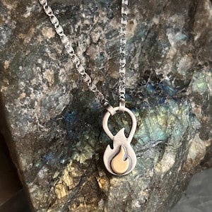 May include: A silver-toned necklace featuring a pendant shaped like a stylized flame with a smaller, smooth, gold-toned flame insert at the center. The pendant hangs from a textured silver chain with repeating star-like patterns along the links.