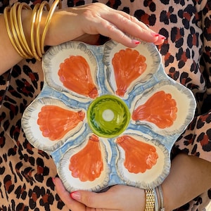 May include: A hand-painted ceramic oyster serving platter. The platter features six oyster shell-shaped compartments in white with orange oyster details, a central green sauce well, and a blue and gray border. A unique piece for serving seafood.