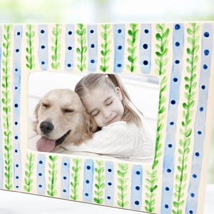 May include: A white picture frame with a blue and green striped pattern. The frame has a photo of a girl hugging a golden retriever.