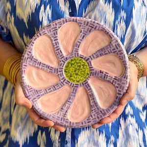 May include: A round, ceramic serving platter with a floral design. The platter features a lavender rim with a repeating pattern, pink petal-shaped sections, and a green center. The platter is held by a person wearing a blue and white patterned shirt.