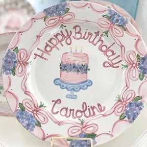 May include: A white ceramic plate with a pink border and pink ribbons. The plate has a hand-painted design of a pink cake with blue frosting and three candles. The text "Happy Birthday Caroline" is written in pink on the plate.