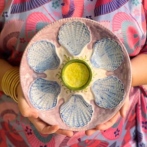 May include: A ceramic oyster serving platter with six shell-shaped compartments and a central well. The platter is light pink with blue and white shell designs. The central well is yellow with a green rim.