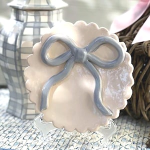 May include: A ceramic trinket dish with a scalloped edge and a large, light blue bow on top. The dish is a pale pink color and is sitting on a clear plastic stand.
