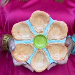 May include: A ceramic oyster serving platter with six oyster shell-shaped compartments and a central round well. The platter is light blue with orange and brown oyster shell details and a green center. The platter is held in front of a pink background.