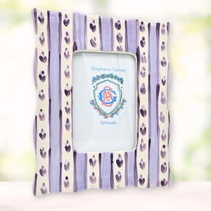 May include: A white ceramic picture frame with a purple and white striped design and a floral pattern. The frame has a scalloped edge and says "Stephanie Garcia Artwork" on the front.