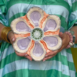 May include: A ceramic oyster serving platter in a flower shape. The platter has six oyster shell-shaped compartments in lavender, surrounded by a peach-colored border. The center features a small, round well with a green wreath design.