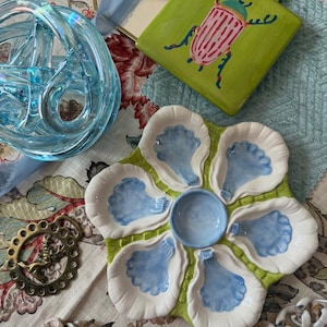 May include: A collection of decorative items including a blue glass knot, a green square tile with a pink and black beetle design, and a flower-shaped oyster serving dish with blue and white accents. A gold-colored decorative ring and a strand of white beads are also present.