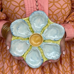 May include: A ceramic oyster serving platter with six oyster shell-shaped compartments and a central dipping well. The platter features a light blue and yellow color scheme, with a glossy finish. The platter is held in front of a pink and orange background.