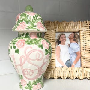 May include: A decorative ceramic jar with a lid, featuring a floral design of pink roses and green leaves. The jar has a pink monogram and is placed next to a woven picture frame with a photo.