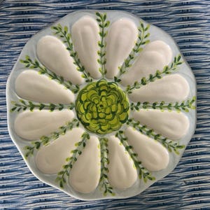 May include: A round ceramic oyster plate featuring twelve recessed white oval wells arranged in a radial pattern. The center is decorated with a vibrant green painted floral motif, while delicate green vine accents separate each white well.
