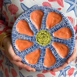 May include: A round, blue and white ceramic oyster serving platter with eight orange oyster-shaped wells and a green center. The platter is held by a person wearing a white top with red and blue starfish and seashell patterns.