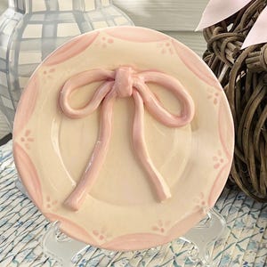 May include: A decorative ceramic plate with a raised pink bow design. The plate has a scalloped edge and a white background with pink accents.