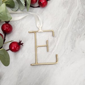 May include: Gold letter E ornament with a white ribbon. The ornament is on a white marble background with a sprig of greenery and red berries.