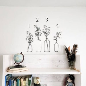 May include: A white wall with four wire art plant designs numbered 1 through 4. Below, a white shelf displays a globe, books, and a vase of feathers. The shelf is against a white wall.