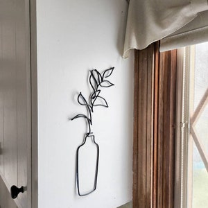 May include: A black metal wire sculpture of a vase with leaves and a stem, mounted on a white wall. The minimalist design features a simple outline of a vase holding a plant. The sculpture adds a touch of modern art to the room.