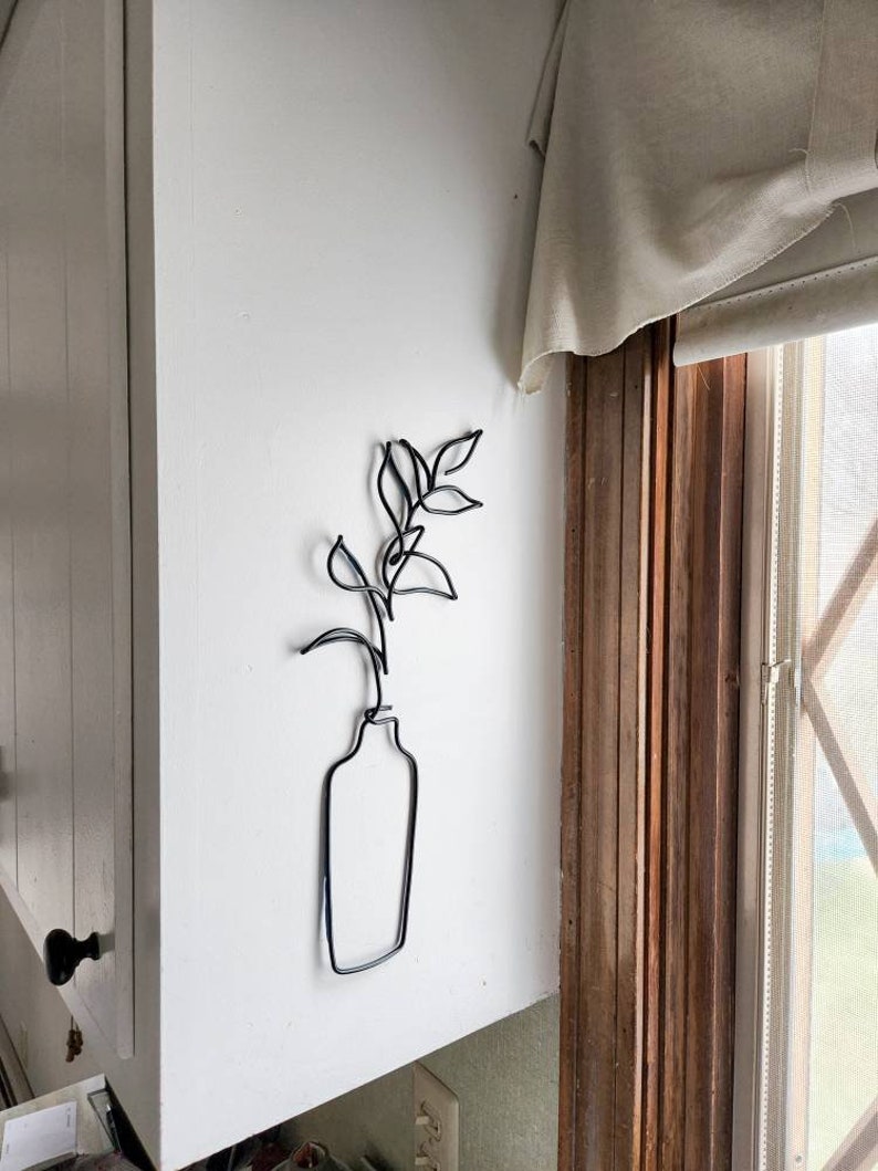 May include: A black metal wire sculpture of a vase with leaves and a stem, mounted on a white wall. The minimalist design features a simple outline of a vase holding a plant. The sculpture adds a touch of modern art to the room.