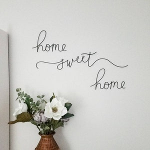 May include: A white wall with black wire lettering that reads "home sweet home". A vase with white flowers and green leaves sits on a surface below the lettering.