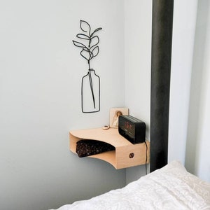 May include: A minimalist bedroom scene features a light wood floating nightstand with a curved design. A black digital alarm clock sits on the nightstand. A black wire art piece of a plant in a vase hangs on the wall. A bed with white bedding is in the foreground.