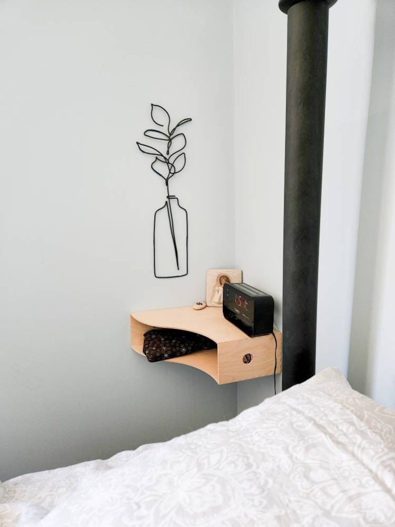 May include: A minimalist bedroom scene features a light wood floating nightstand with a curved design. A black digital alarm clock sits on the nightstand. A black wire art piece of a plant in a vase hangs on the wall. A bed with white bedding is in the foreground.