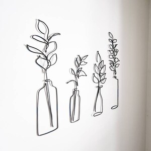 May include: Four black wire art pieces depicting stylized plants in vases, mounted on a white wall. The pieces vary in size and shape, with the leaves and vases outlined in a continuous wire form. The minimalist design adds a modern touch to the decor.
