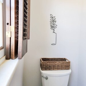 May include: A white toilet with a woven basket on top. A black metal wall art piece of a plant is mounted on the wall. A wooden shutter is partially open next to the toilet.