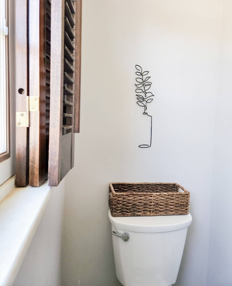 May include: A white toilet with a woven basket on top. A black metal wall art piece of a plant is mounted on the wall. A wooden shutter is partially open next to the toilet.