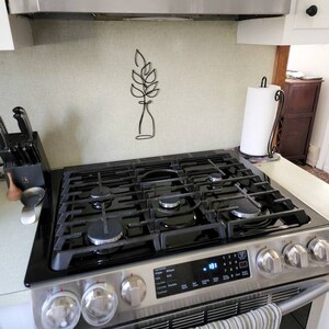 May include: Stainless steel gas range with black grates and control panel. A striped towel hangs from the handle. A black metal wall art piece of a plant is mounted on the wall above the range.