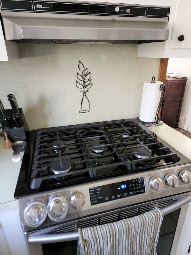 May include: Stainless steel gas range with black grates and control panel. A striped towel hangs from the handle. A black metal wall art piece of a plant is mounted on the wall above the range.