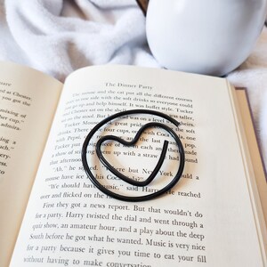 May include: A black metal bookmark shaped like a heart is placed on an open book. The book is titled "The Dinner Party" and the text on the page discusses a dinner party with a buffet style.