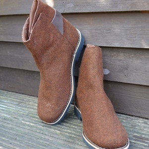 May include: A pair of brown felt boots with black soles and a brown leather strap on the back of the boot.