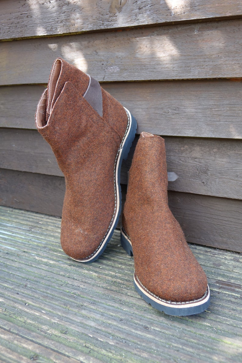 May include: A pair of brown felt boots with black soles and a brown leather strap on the back of the boot.