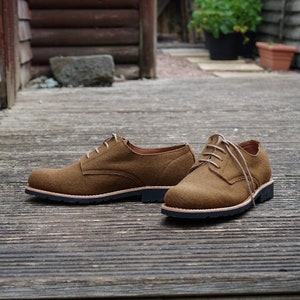 May include: A pair of brown suede lace-up shoes with a dark brown sole. The shoes are on a wooden deck.