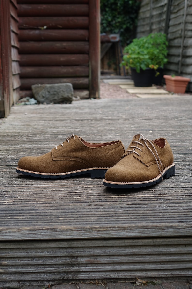 May include: A pair of brown suede lace-up shoes with a dark brown sole. The shoes are on a wooden deck.